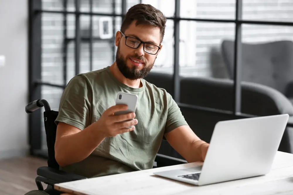 The image shows a soldier in a wheelchair using a mobile phone at home. This image represents the context of ADA (Americans with Disabilities Act) Remediation, emphasizing the importance of ensuring accessibility for individuals with disabilities, even in their own homes.