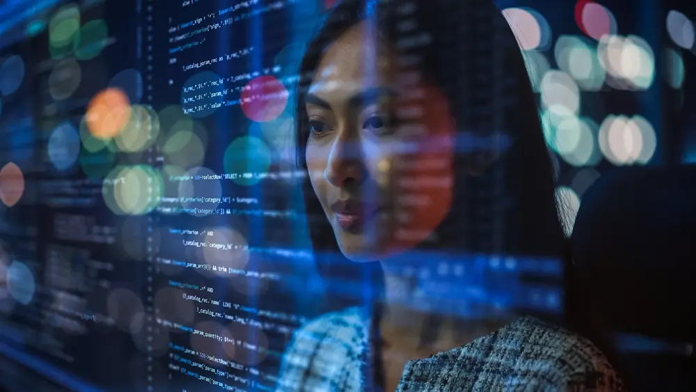 A portrait of an Asian female startup digital entrepreneur is shown working on her computer, with a line of code projected on her face and reflecting her dedication. She is depicted as a software developer working on an innovative e-commerce AI app, emphasizing big data. This image highlights the context of Accessibility Remediation, showcasing technology's potential to empower inclusive digital ventures.