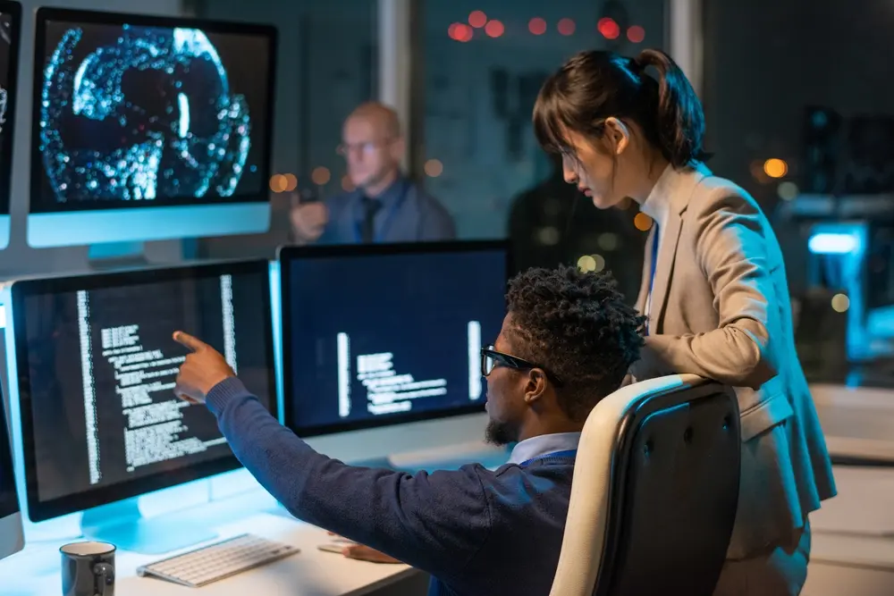 In a meeting, a confident businessman is shown pointing at coded data on a computer screen while explaining it to his female colleague. This image represents the context of Automation Consulting Services, highlighting strategic guidance and collaboration in implementing automation solutions.