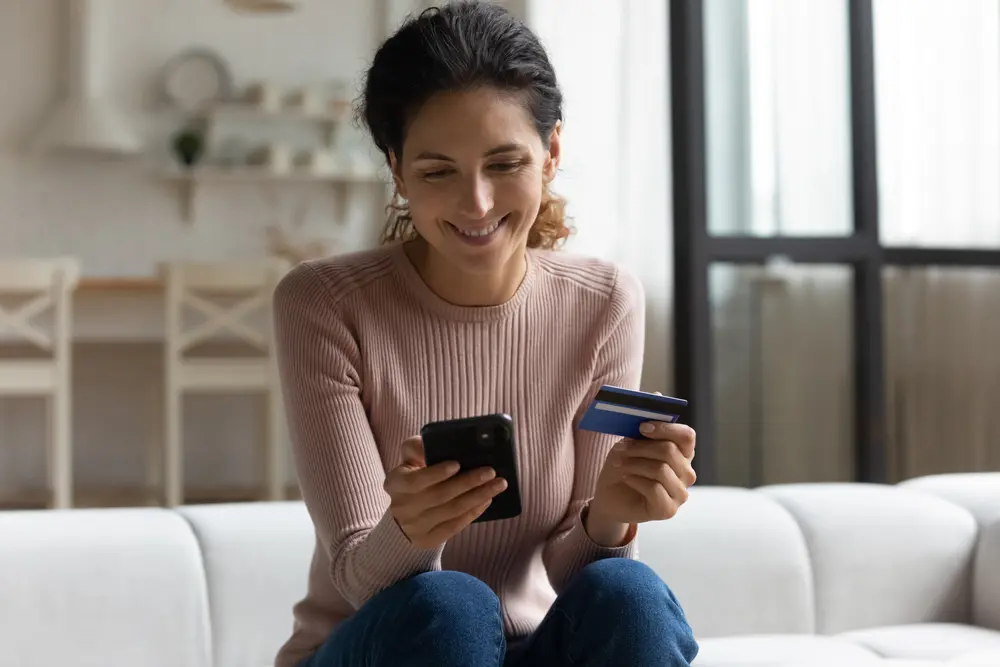 A smiling Latina lady client is depicted holding a mobile phone and a credit bank card while doing online shopping and providing internet payment. This happy young woman is enjoying easy, fast, and secure money transfers in a banking app. This image represents the context of Banking Software Testing, emphasizing safe and secure mobile banking transactions.