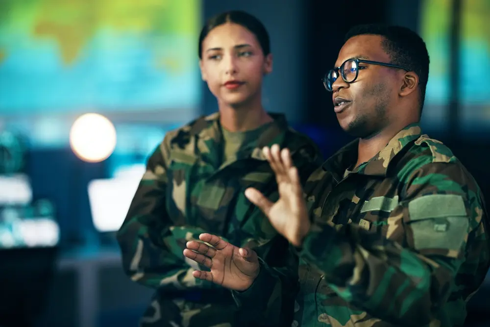 In a nighttime scene, a diverse group of people in the military is engaged in a strategic discussion related to cybersecurity or military communication. A black man is shown speaking to a woman about government secrets, surveillance, or system operations. This image represents the context of a CMMC Consultant providing guidance and advice to enhance military cybersecurity readiness and compliance.