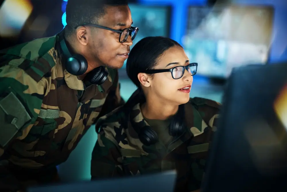 In a military setting, employees are seen collaborating on a project related to cybersecurity, communication, or control room monitoring. The scene depicts a group of people with computers discussing strategies in an office environment. This image represents the context of CMMC Readiness Assessment, highlighting the military's efforts to prepare for compliance with cybersecurity standards.
