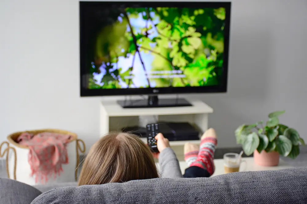 A young woman sits comfortably on a sofa in her living room, watching television with subtitles on the screen. The serene atmosphere of nature and greenery adds to the ambiance, highlighting the importance of Closed Captioning Services in ensuring accessibility for all viewers.