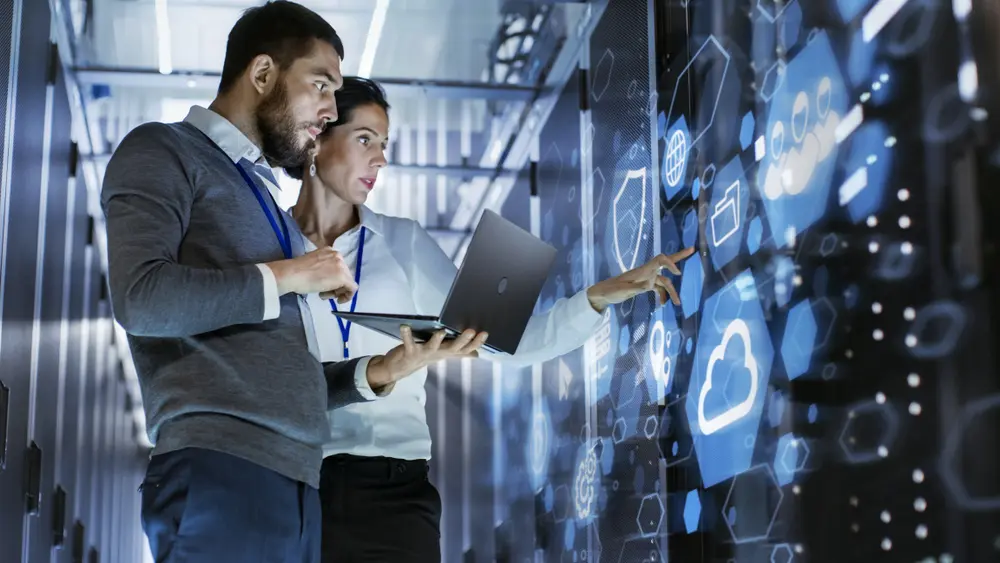 A male IT specialist holds a laptop and discusses work with a female server technician in a data center. Behind them is a rack server cabinet with a cloud server icon and visualization. This image contextualizes Cloud Assessment Services, illustrating professionals' collaboration in ensuring secure and efficient cloud infrastructure.