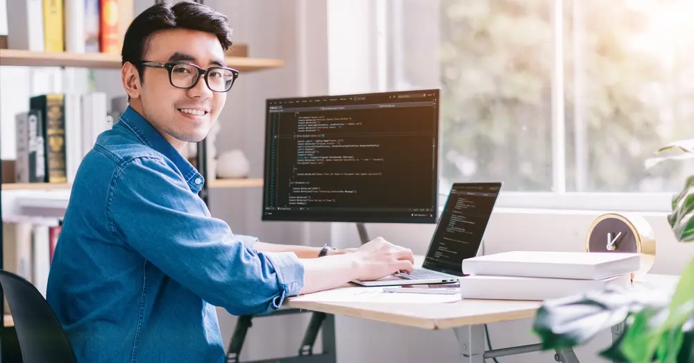 In an office setting, a young Asian programmer is shown focused on his work. The context is Compatibility Testing Services, highlighting his role in ensuring software compatibility across different platforms and environments.