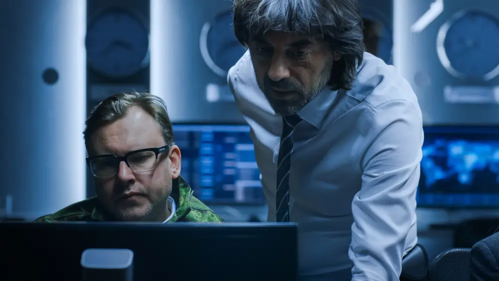 In a system control room, the Government Chief of Cyber Security consults with an Operations Officer who is working on a computer. Specialists are seen working on computers in the background. This image represents the context of Compliance Consulting Services, highlighting the importance of ensuring regulatory compliance in cybersecurity practices.