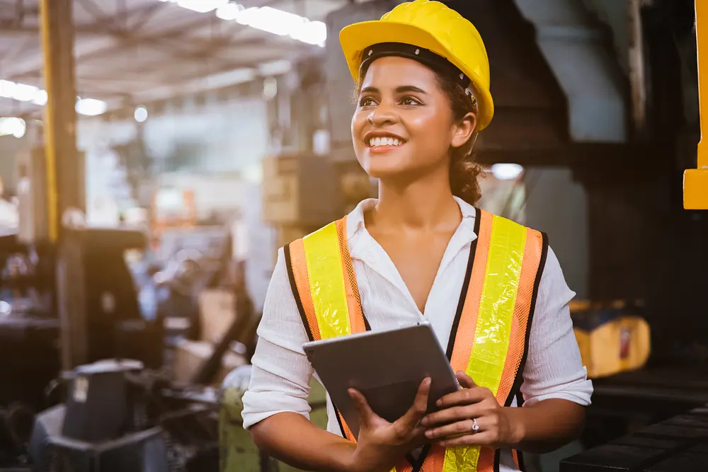 A dark-skinned industry maintenance engineer woman, wearing a uniform and safety helmet, is shown under inspection and checking the production process on a factory station using a tablet. This image represents the context of Construction Software Testing, emphasizing the importance of ensuring integrity and reliability in construction software used for production processes.