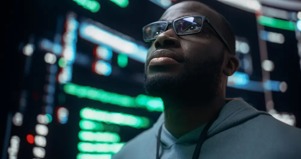 In a close-up portrait, a young Black man is shown looking at big digital screens glitching while displaying code. He appears worried as he works on fixing a bug, dealing with a crashing system, and thinking of solutions. This image represents the context of Cyber Security Assessment Service, highlighting the diligent efforts of professionals in securing digital systems.