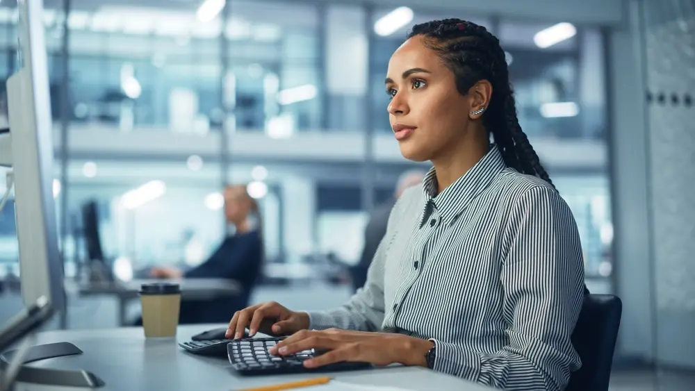 In a corporate office setting, a black female IT technician is seen using a desktop computer, working with big data statistics and graphs. Meanwhile, a smiling creative software engineer is shown working on an eCommerce project marketing and development. This image represents the context of DevOps Testing Services, illustrating collaboration and innovation in the testing process.