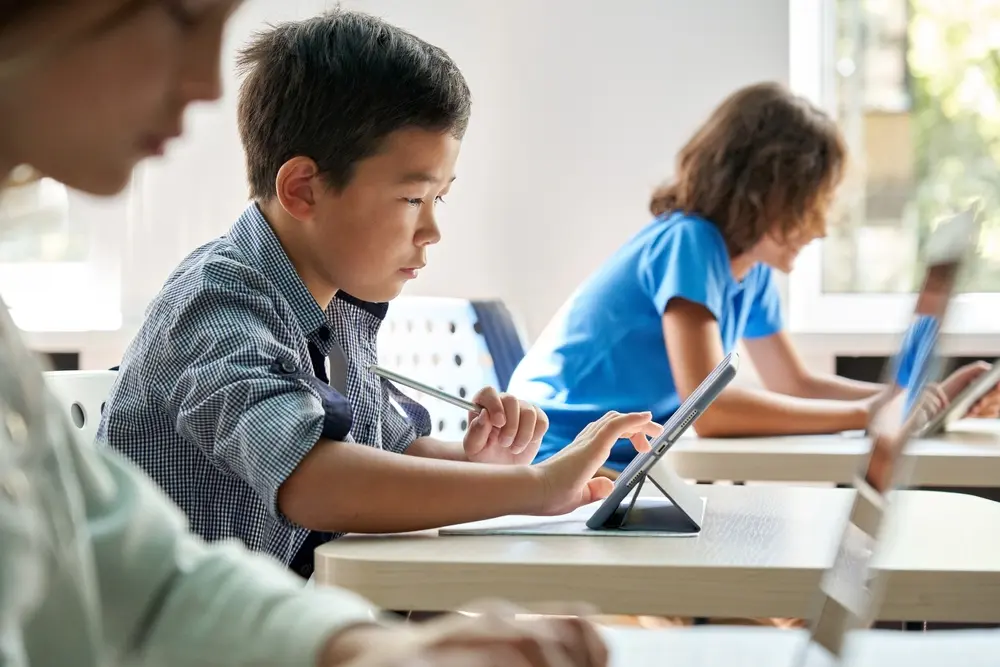 In a classroom setting, a focused Asian school boy is seen using a digital tablet. He is an attentive junior school student learning online through a virtual education digital program app during a STEM computer science lesson. This image represents the context of Educational Software Testing, highlighting the importance of testing software used in educational environments for effective online learning experiences.