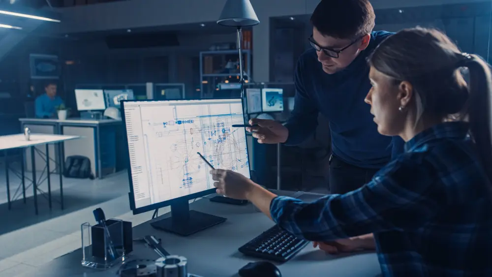 In an industrial design engineering facility office, an engineer is working on a desktop computer, with the screen displaying CAD software and technical blueprints. Her male project manager explains job specifics, emphasizing collaboration and innovation. This image contextualizes Exploratory Testing Services, showcasing the team's dedication to exploring new ideas and solutions.