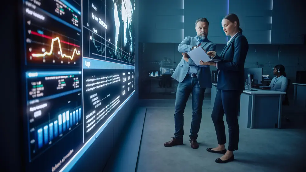 In a telecommunications company's system control and monitoring room, a project manager and a computer science engineer are engaged in discussion while using a big screen display and a laptop. They are showing infrastructure infographics data. This image represents the context of ISO 27001 Consulting Services, highlighting collaboration and expertise in information security management.
