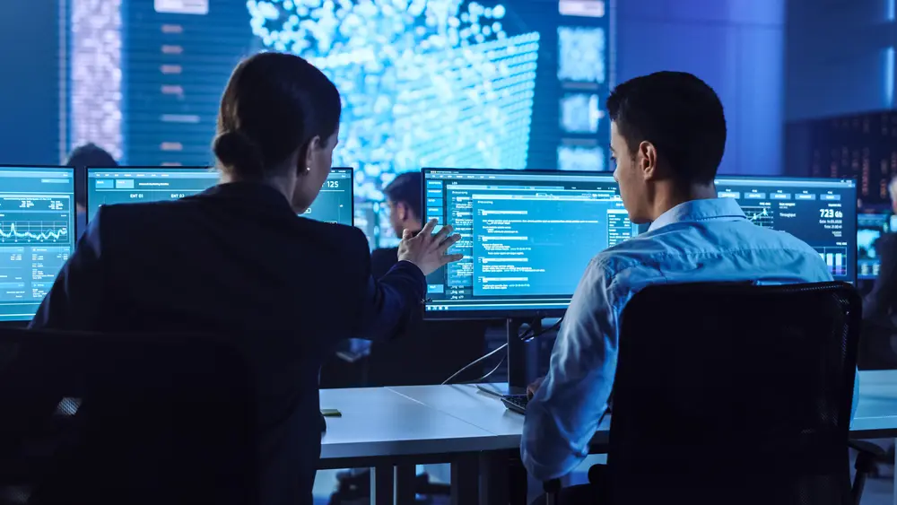 In a big control and monitoring room with a neural network, a confident female data scientist works closely with her young colleague. Young specialists are seen talking and discussing work in a room with other employees. This image represents the context of ISO 27001 Gap Analysis, emphasizing the collaboration and expertise involved in identifying security gaps and ensuring compliance with information security standards.