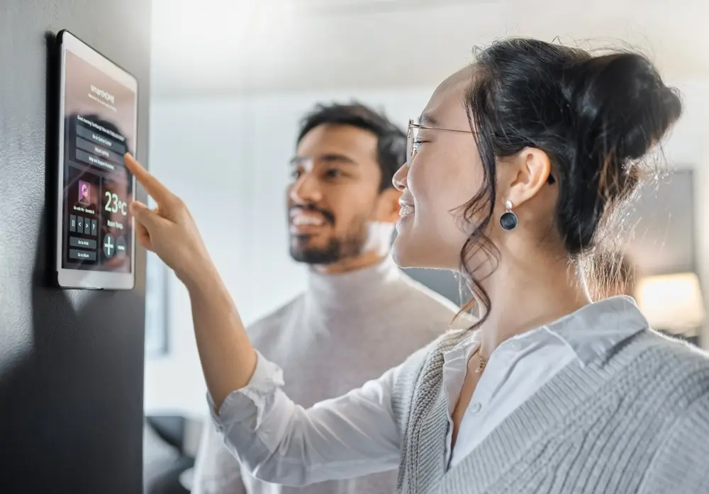A couple is depicted interacting with smart home technology, including a wall system and digital dashboard for air conditioning, safety security network, and house automation. They are seen using an app software and UI panel to program IoT monitoring devices. This image represents the context of IoT Testing Services, emphasizing the integration and testing of interconnected devices for seamless home automation.