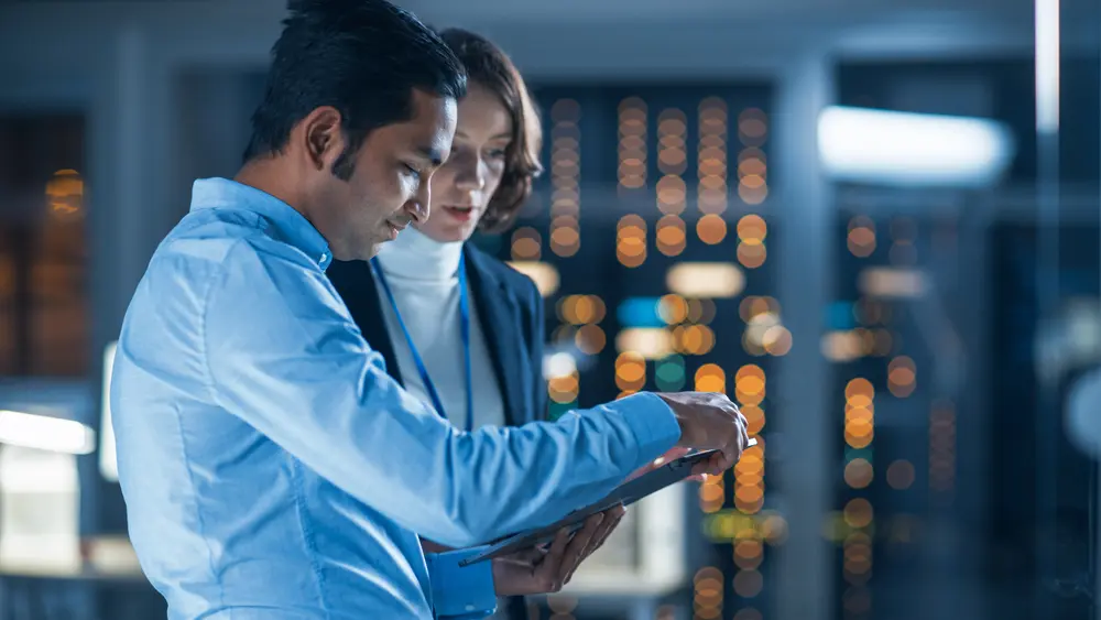 In a technology research facility, a female project manager is seen consulting with the chief engineer as they discuss information on a tablet computer. Meanwhile, a team of industrial engineers and developers are working on engine design using computers. This image embodies the context of a NIST 800-171 Consultant, emphasizing collaboration and expertise in navigating compliance within the technology research environment.