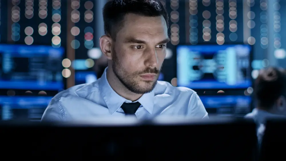 A close-up of a professional technical controller sitting at his desk with multiple displays before him. In the background, his colleagues are working in the system control center. This image represents the context of NIST 800-171 Gap Assessment, highlighting the meticulous evaluation of security controls and standards in the system control environment.