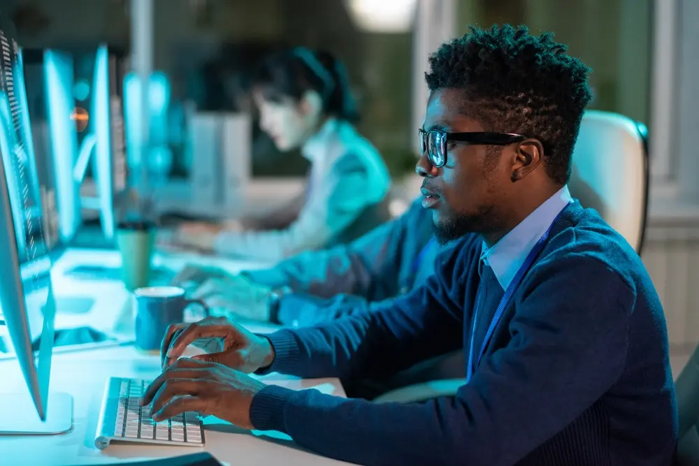 A young serious businessman in eyeglasses and formalwear is depicted looking at the screen of a computer, with his coworkers in the background. This image represents the context of a Penetration Testing Report, illustrating the careful analysis and consideration of security vulnerabilities within the organization.