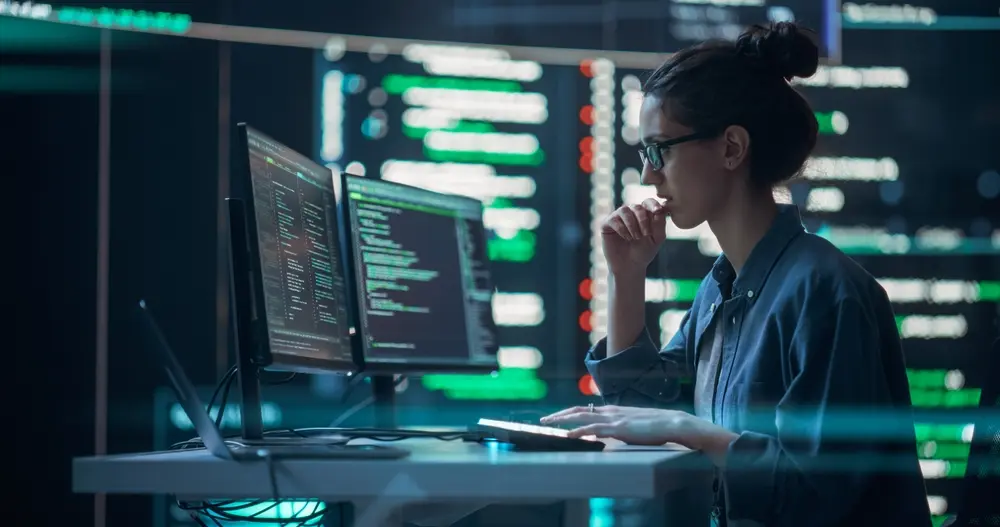A female developer is shown thinking and typing on a computer, surrounded by big screens displaying coding language. She is a focused programmer creating software and running coding tests, embodying a futuristic concept of programming. This image represents the context of Penetration Testing Services, highlighting the meticulous approach to testing software security vulnerabilities.
