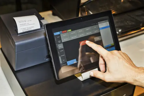 In a coffee cafe, a waiter is shown holding a POS terminal while serving customers and touching the screen of a tablet with software interface to take orders and print receipts. This image represents the context of Retail Software Testing, emphasizing the importance of testing software used in retail environments for efficient order processing.