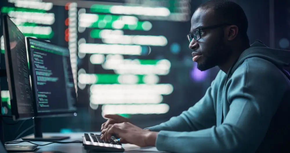 A black male programmer is depicted working in a monitoring room surrounded by big screens displaying lines of programming language code. This portrait showcases him creating software, embodying a futuristic coding concept. The image represents the context of Risk Assessment Services, highlighting proactive efforts to secure digital systems.