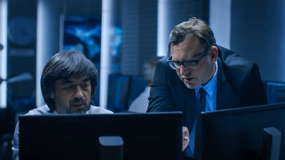 In a system control room, the Government Chief of Cyber Security is consulting with an Operations Officer who is working on a computer. Specialists are seen working on computers in the background. This image represents the context of Risk Management Consulting Services, illustrating collaboration and expertise in managing cybersecurity risks at the government level.