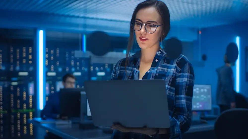 In the foreground, a smart and seductive young woman wearing glasses holds a laptop. In the background, there is a technical department office with specialists working and functional data server racks. This image represents the context of a Security Assessment Report, highlighting the expertise and diligence of professionals in assessing security measures.