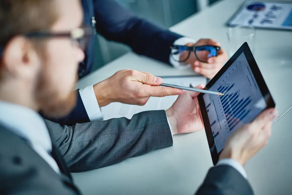 A businessman is holding a touchpad while his colleague explains data to him. This image represents the context of Software Performance Testing Services, illustrating the collaborative effort to analyze and improve software performance.