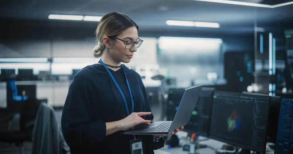 In a room with a big green screen mock-up display, a multiethnic QA engineer is shown working on finding and fixing bugs in a product or software before launch. She is a female using a laptop, illustrating her expertise in stress testing services.