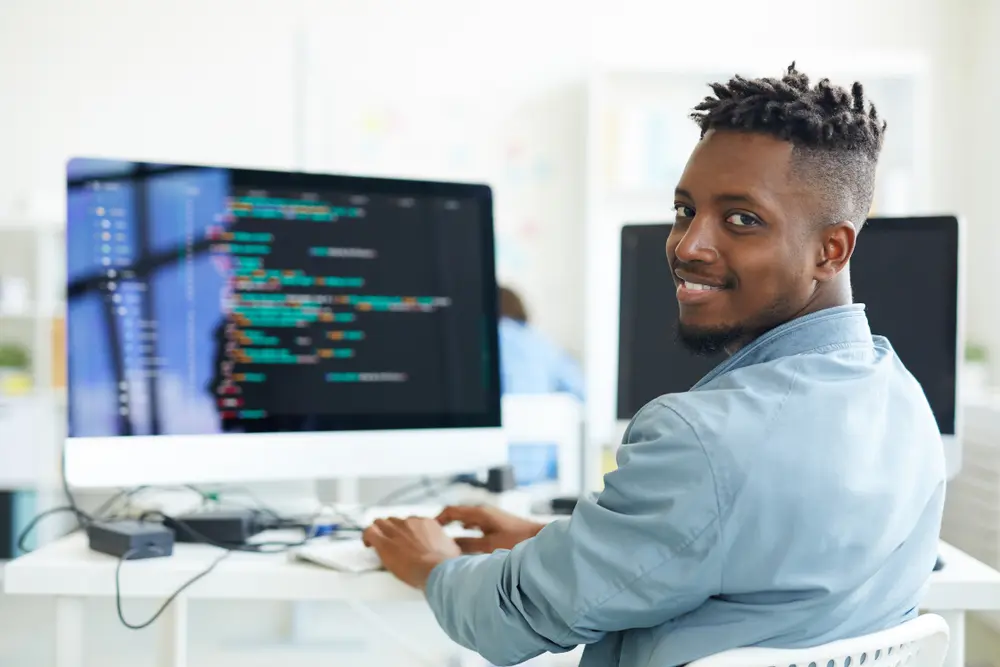 A happy guy is shown looking at you while sitting by his workplace and working with software. This image represents the context of Test Automation Services, showcasing his efficient work in automating tests.