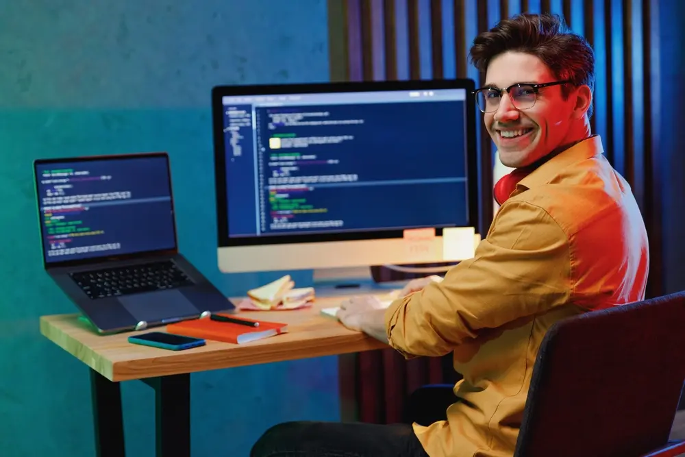 A smiling young full-stack developer, software engineer, IT specialist, and programmer man in a shirt is shown working at home, writing code on a laptop PC computer and checking the database. This image represents the concept of program development and is contextualized within Usability Testing Services, emphasizing the importance of ensuring user-friendly software interfaces.