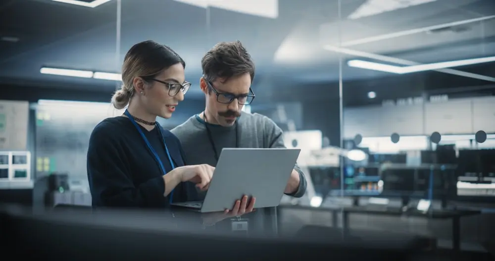 The image shows two creative young female and male engineers using a laptop computer to analyze and discuss how to proceed with artificial intelligence software. They are standing in a high-tech research office. This scene represents the context of WCAG (Web Content Accessibility Guidelines) Consulting, highlighting efforts to ensure accessibility in advanced technological developments.
