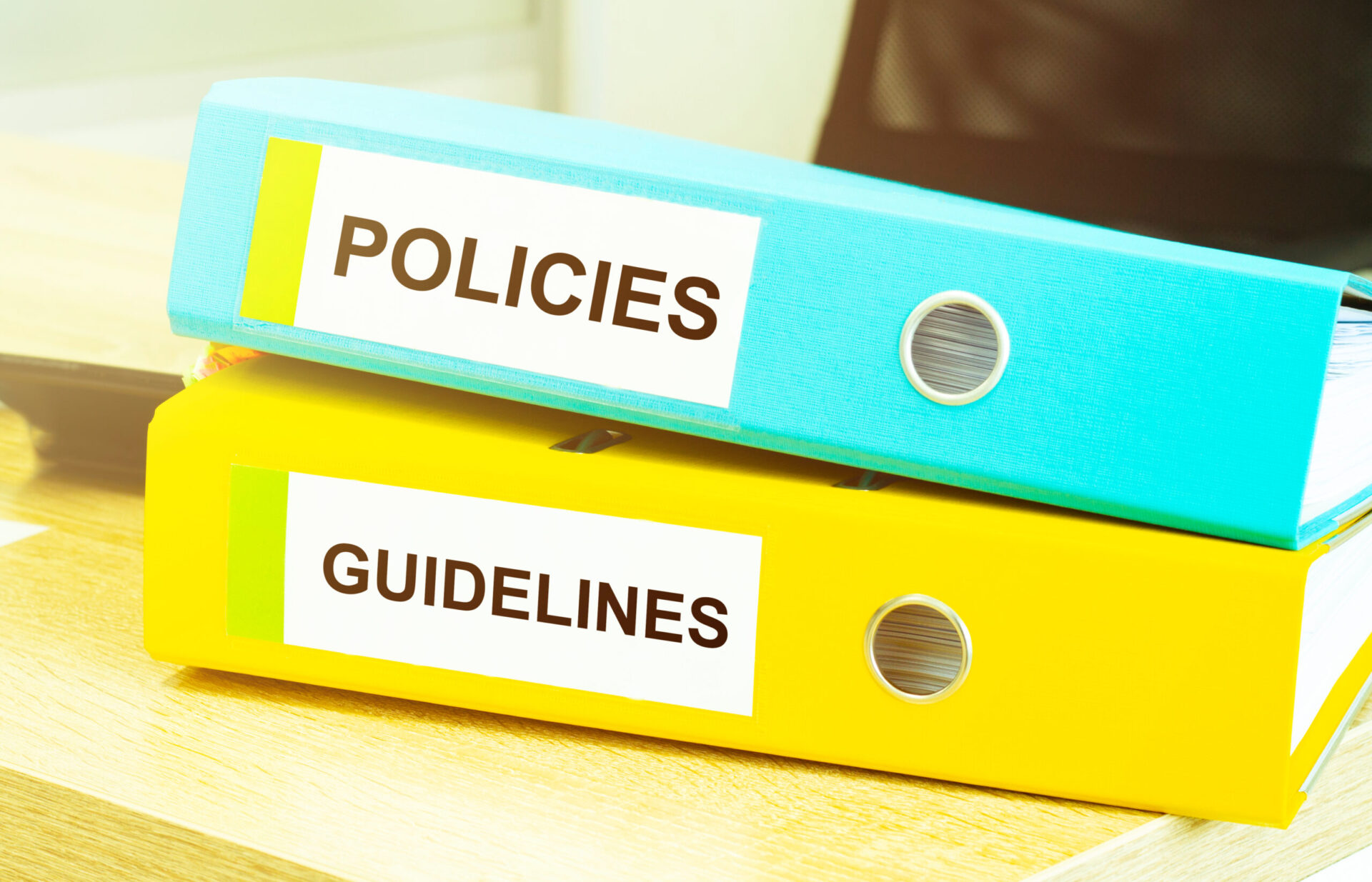 A blue binder that reads "Policies", lays over a yellow binder that reads "Guidelines". Both binders sit on top of a desk.