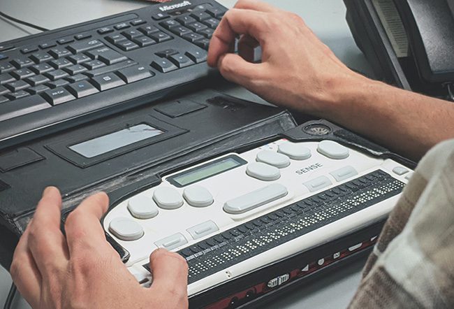A Tester's hands on a Braille display beside a keyboard during a manual accessibility assessment
