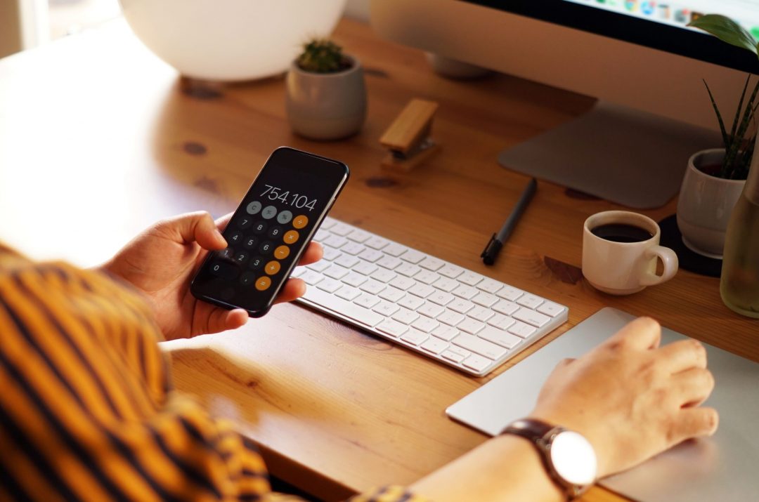 A person holding a smartphone displaying a calculator app with a result of 754.104. They are seated at a wooden desk with a keyboard, touchpad, and a small cup of coffee in the background, suggesting a productive work environment.