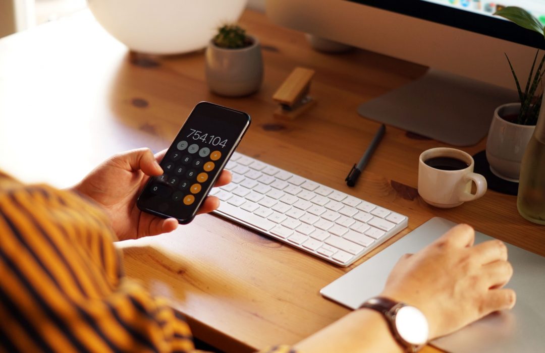 A person holding a smartphone displaying a calculator app with a result of 754.104. They are seated at a wooden desk with a keyboard, touchpad, and a small cup of coffee in the background, suggesting a productive work environment.