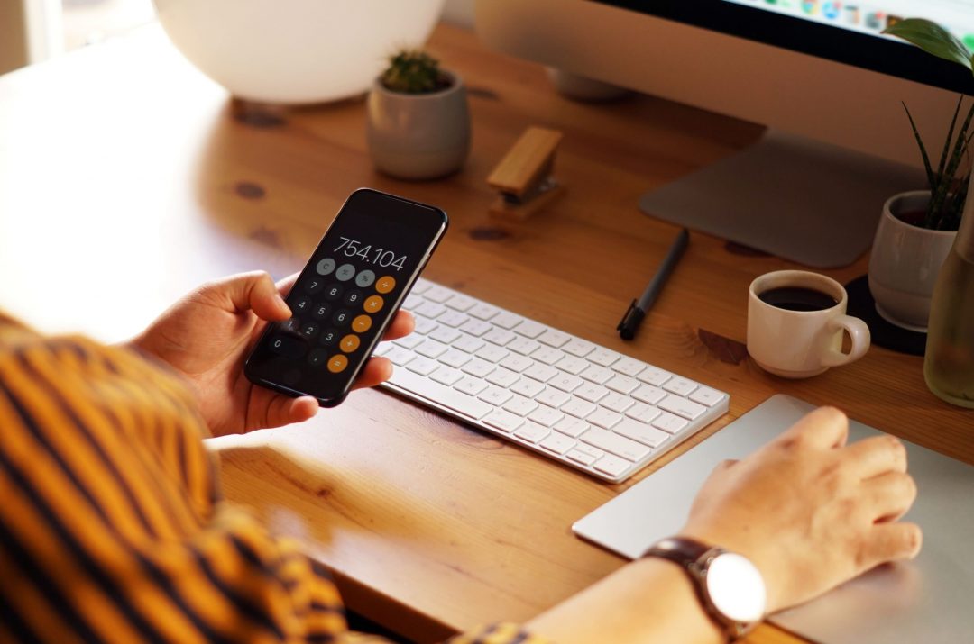 closeup-shot-entrepreneur-working-from-home-doing-calculations-min A person holding a smartphone displaying a calculator app with a result of 754.104. They are seated at a wooden desk with a keyboard, touchpad, and a small cup of coffee in the background, suggesting a productive work environment.
