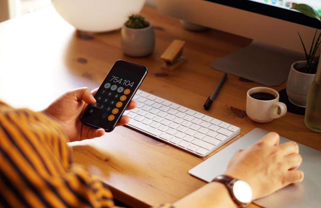 closeup-shot-entrepreneur-working-from-home-doing-calculations-min A person holding a smartphone displaying a calculator app with a result of 754.104. They are seated at a wooden desk with a keyboard, touchpad, and a small cup of coffee in the background, suggesting a productive work environment.