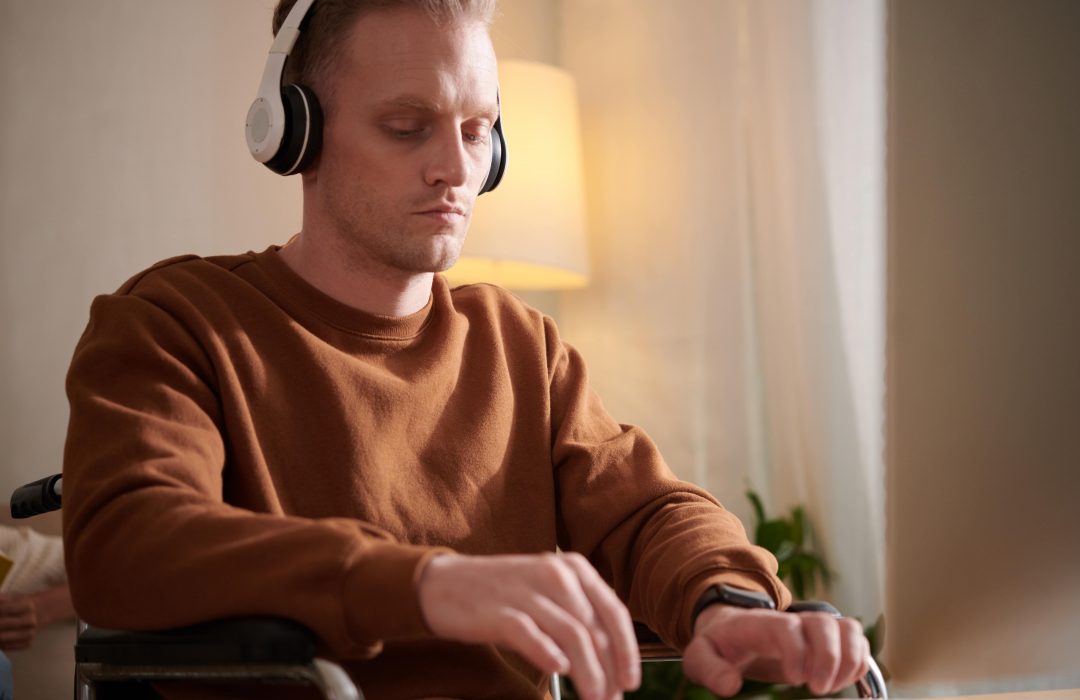 A man in a wheelchair wearing headphones and a brown sweatshirt is using a computer or touchscreen device, appearing focused in a warmly lit indoor setting.