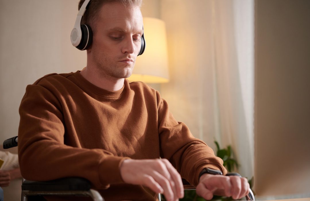 man-with-disability-working-computer-min A man in a wheelchair wearing headphones and a brown sweatshirt is using a computer or touchscreen device, appearing focused in a warmly lit indoor setting.
