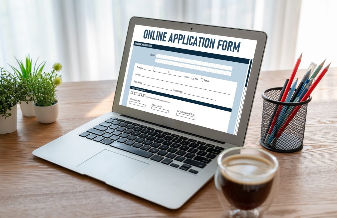 A laptop on a wooden desk displaying an online application form with fields for personal information, including name, birth date, address, phone number, email, and ID details. A cup of coffee and a container of pens are beside the laptop, emphasizing a work or registration setting. The form highlights the importance of digital accessibility for users relying on assistive technologies.