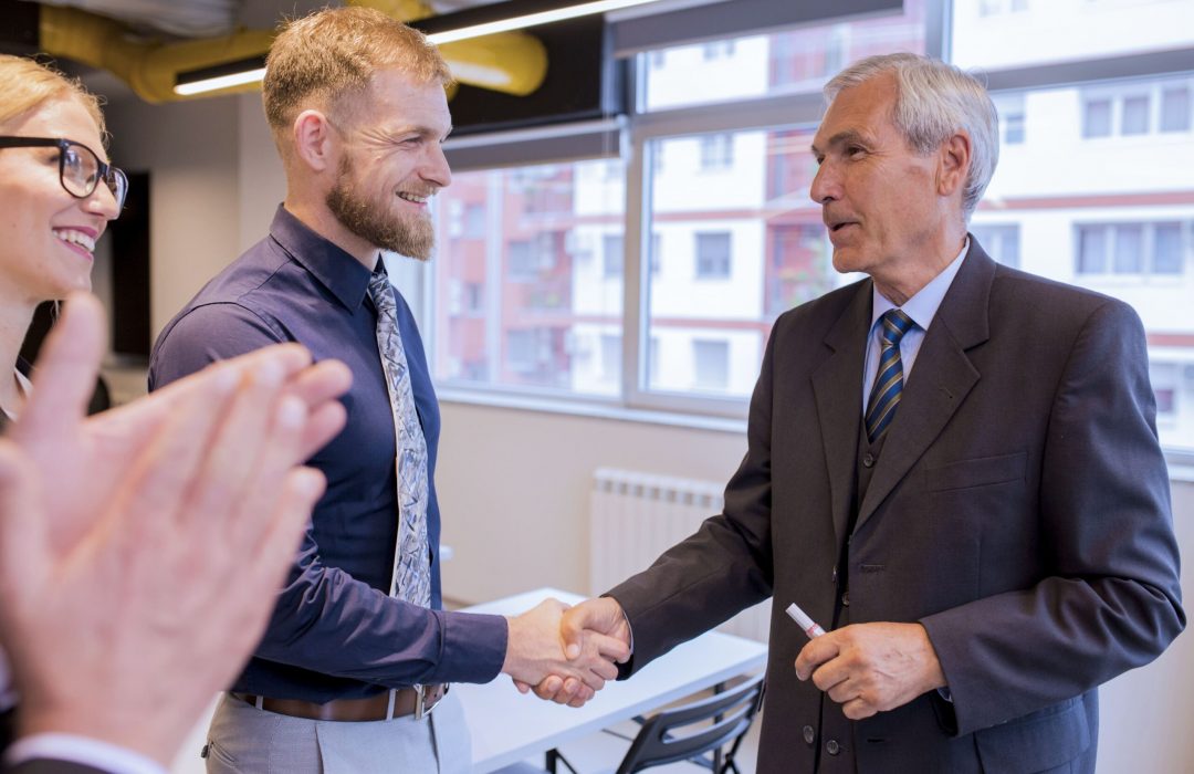 A professional setting where two men in business attire are shaking hands. One man is elderly and wearing a suit, while the younger man is dressed in a shirt and tie. They are smiling, and other colleagues are applauding in the background.