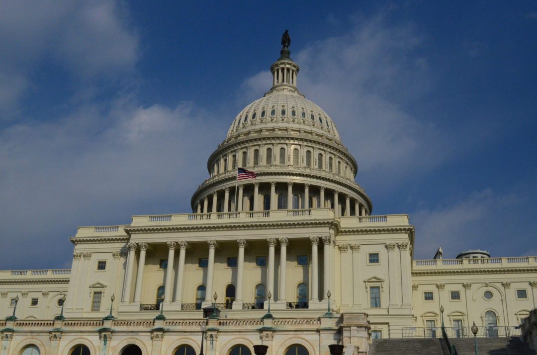 The United States Capitol Building in Washington, D.C., with an American flag flying in front of its dome against a blue sky with scattered clouds.